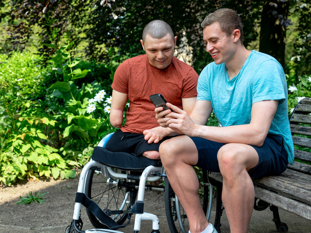 A young man who is a wheelchair user looking at a mobile phone with a friend in the park
